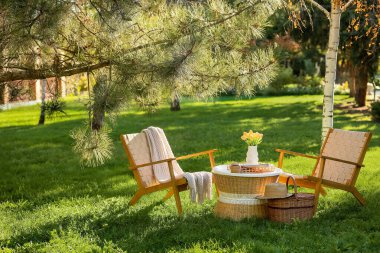 Sepeti, dergileri ve vazosu olan rahat hasır koltuklar parktaki kahve masasında, açık havada.