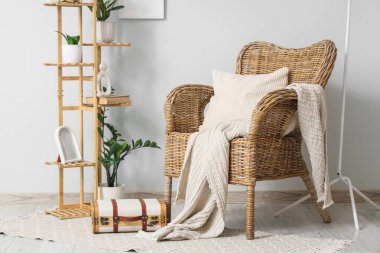 Hasır koltuklu, çiçekli, raflı ve bavullu oda. Beyaz duvarın yanında.