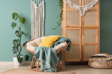 Koltuk, ev bitkileri, dolap, hasır döşeme ve mavi duvarda dokuma halısı olan odanın içi.