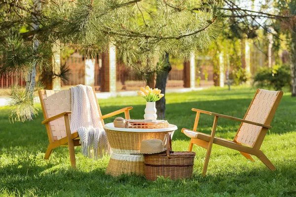 Sepeti, dergileri ve vazosu olan rahat hasır koltuklar parktaki kahve masasında, açık havada.