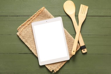 Yeşil ahşap arka planda peçete ve mutfak aletleri olan dijital tarif kitabı.