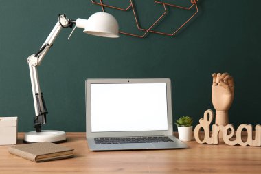 Yeşil duvarın yanındaki masada lamba olan boş bir dizüstü bilgisayar.