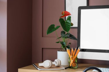 Ofisteki kahverengi duvarın yanında bilgisayar, kulaklık ve anthurium çiçekli modern işyeri