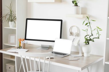 Ofiste beyaz duvarın yanında bilgisayar, dizüstü bilgisayar ve kulaklık bulunan modern işyeri