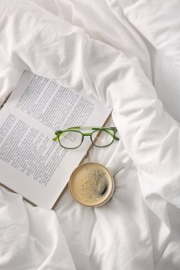 Beyaz çarşafların üzerinde kitap ve gözlüklü bir fincan kahve.