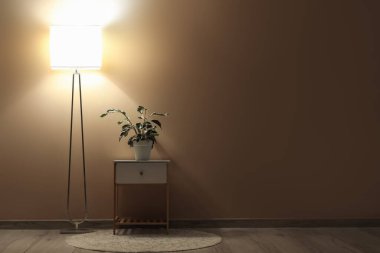 Beyaz komodinli, çiçekli ve bej duvarın yanında parlayan bir lamba.