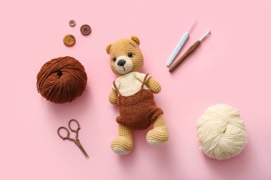 İplikli ayı oyuncağı ve pembe arka planda örgü aletleri.