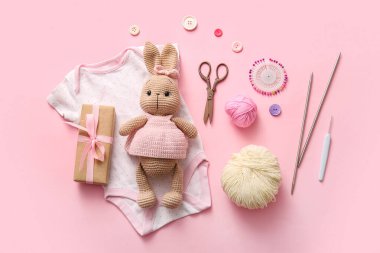 Örgü aletleri, bebek kıyafetleri ve pembe arka planı olan örülü tavşan oyuncağı.