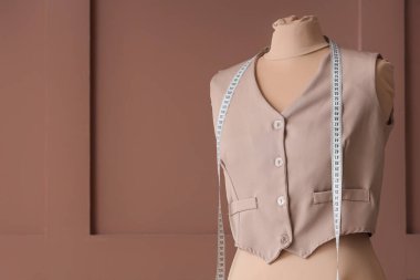 Kahverengi ahşap arka planda yeleği ve mezurası olan terzi mankeni.