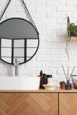 Lavabo kasesi ve banyo aksesuarları beyaz tuğla duvarın yanındaki ahşap dolapta.