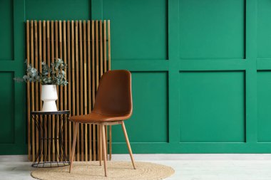 Odanın ahşap perdesinin yanındaki masada okaliptüs dalları olan modern kahverengi sandalye ve vazo.