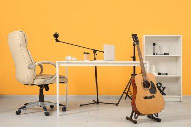 Kayıt stüdyosundaki sarı duvarın yanında dizüstü bilgisayarı, kahve fincanı ve müzik ekipmanı olan bir iş yeri.