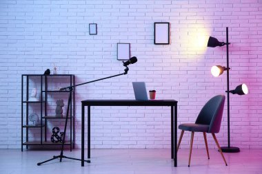 Kayıt stüdyosundaki beyaz tuğla duvarın yanında dizüstü bilgisayarı, kahve fincanı ve mikrofonu olan bir iş yeri.