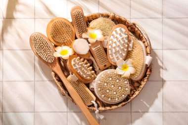 Farklı masaj fırçaları olan sepet ve beyaz karo arka planda plumeria çiçekleri