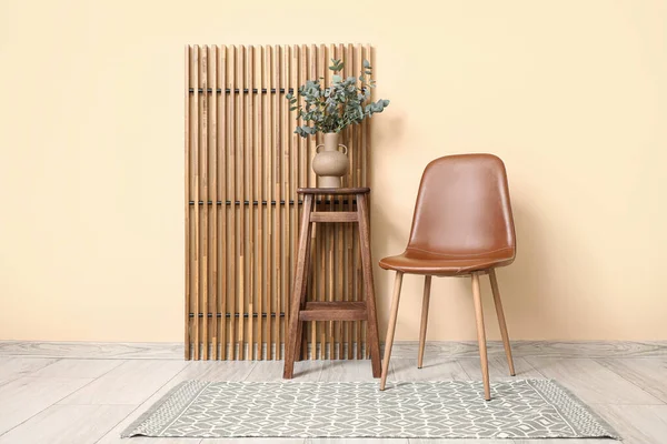 Odanın ahşap perdesinin yanındaki taburede okaliptüs dalları olan modern kahverengi sandalye ve vazo.