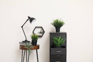 Masa lambası küçük masada ve dosya dolabında beyaz duvarın yanında bitkiler var