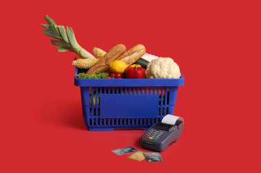 Ödeme terminali ve kredi kartlarıyla dolu alışveriş sepeti kırmızı arka planda.