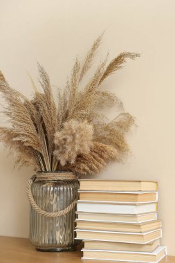 Bej duvarın yanındaki rafta otlu vazo ve kitap yığını.