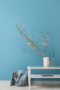 Mavi duvarın yanındaki cam sehpanın üzerinde çiçek açan dalları, mumları ve dizüstü bilgisayarı olan vazo.