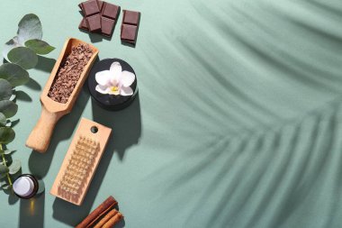 Yeşil arka planda ahşap kepçe çikolata, yeşil dal ve bir kase kozmetik ürünü içeren spa kompozisyonu