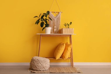 Sarı duvarın yanında bitki, yastık ve puf dolu bir masa.