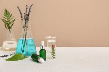 Bej duvarın yanındaki beyaz masada doğal kozmetik ürünleri hazırlamak için malzemeler ve numunelerle dolu laboratuvar vitrinleri.