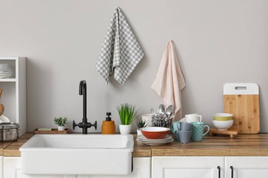 Ahşap mutfak tezgahı, temiz tabaklar, seramik lavabo ve gri duvarda asılı havlular.