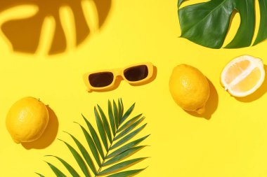 Sarı arka planda güneş gözlüğü, limon ve tropikal yapraklı yaz kompozisyonu