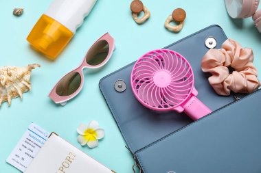 İçinde kadın aksesuarları, pasaport, bilet ve mavi arka planda mini fan olan bir çanta.