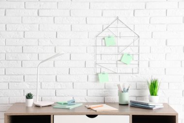 Masa lambası, ev bitkileri ve beyaz tuğla duvarın yanında ofis malzemeleri olan modern işyeri.