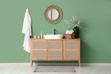 Banyonun içinde, yeşil duvarın yanındaki çekmecelerin üzerinde çiçek açan bir vazo var.