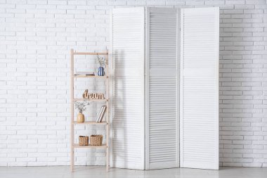 Beyaz tuğla duvarın yanındaki raf ünitesinde katlanır ekran ve çiçek açan ağaç dalları