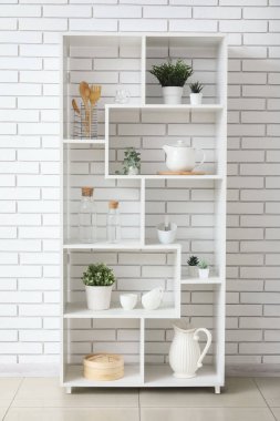 Mutfak aletleri ve beyaz tuğla duvarın yanında yapay bitkiler olan raf ünitesi.