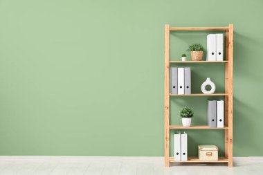 Ofiste yeşil duvarın yanında klasörleri ve yapay bitkileri olan raf ünitesi.