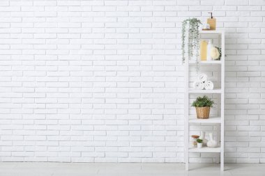 Banyo malzemeleri ve beyaz tuğla duvarın yanında yapay bitkiler olan raf ünitesi.