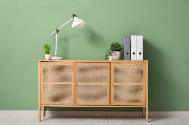 Ofisteki yeşil duvarın yakınındaki çekmecelerin üzerinde dosyalar ve yapay bitkiler olan lamba.