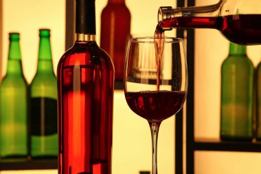 Barda şişeler olan rafların yanındaki bardaklara kırmızı şarap dökülüyor.