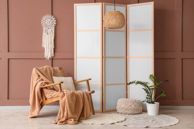 Katlanan ekran, puf, koltuk ve kahverengi duvarın yanındaki saksı.