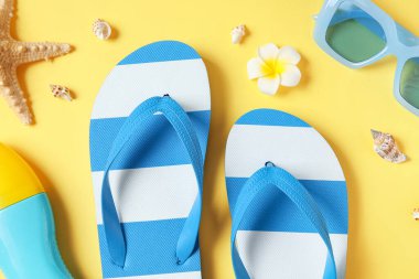 Çiçekli mavi çizgili terlikler, deniz kabukları, bir şişe güneş kremi ve sarı arka planda güneş gözlüğü.