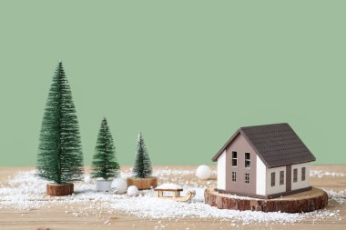 Tahta gövdeli güzel bir ev modeli, dekoratif köknar ağaçları ve yeşil duvarın yanındaki kahverengi masada kar.