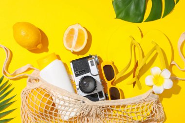 Sarı arka planda sicim torbası, plaj aksesuarları ve tropikal yapraklarla yaz kompozisyonu