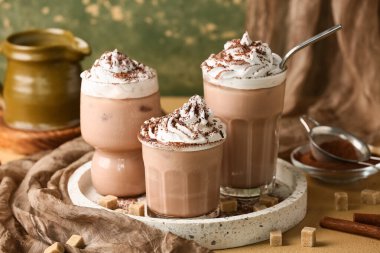 Yeşil grunge duvarına karşı krem şantili, şeker küplü ve tarçın çubuklu kakao bardakları