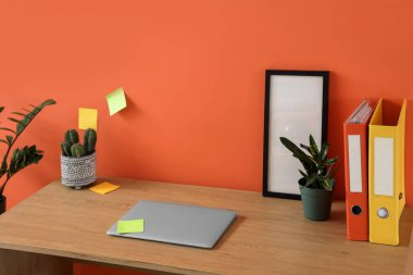 Ofiste turuncu duvarın yanında modern dizüstü bilgisayarı ve ev bitkileriyle rahat bir iş yeri, yakın plan.