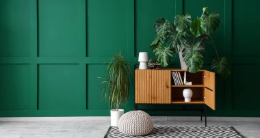 Odanın içinde ahşap klozet ve yeşil duvarın yanında Monstera bitkisi var. Tasarım için pankart