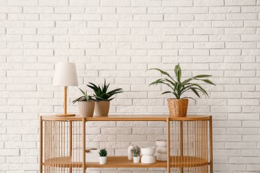 Beyaz tuğla duvarın yanında çiçek ve lamba olan masa.