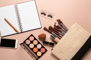 Makyaj ürünleriyle dolu makyaj çantası, modern cep telefonu ve bej arka planda not defteri.