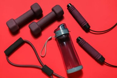 Spor su şişesi, genişletici, dambıl ve kırmızı arka planda atlama ipi.