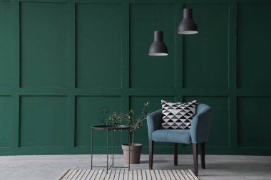 Yastıklı koltuk, sehpa, lamba ve yeşil duvarın yanındaki dekoratif zeytin ağacı.