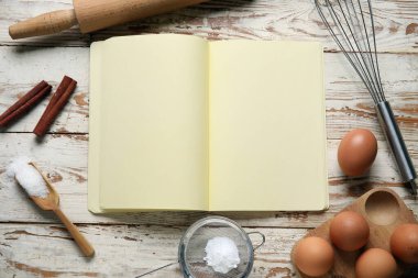 Beyaz ahşap zemin üzerinde boş tarif defteri, yumurta ve kap kacak içeren kompozisyon