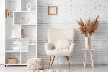 Beyaz tuğla duvarın yanında koltuk, puf ve raf ünitesi olan bir oda.